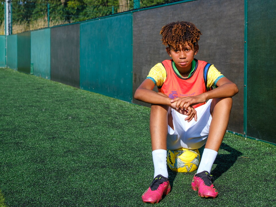 Premier League Kicks participant sits on a football beside a small-sided outdoor pitch, wearing training kit and football boots during a community football session.