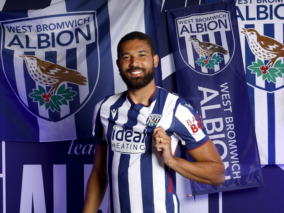 George Campbell in the home kit smiling at the camera and pulling on the badge in front of WBA branding 
